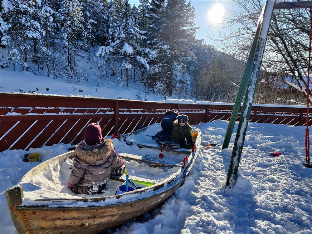 Tre barn sitter i en gammel båt som står ved et lekestativ i snøen.  - Klikk for stort bilde